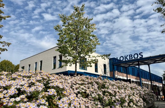 Voorzijde van een gebouw met de naam Blokbos, omringd door bloeiende paarse bloemen en een boom onder een partly cloudy hemel. (vergroot foto)