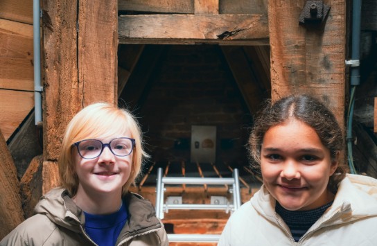 Twee lachende kinderen in warme jassen staan voor een houten zolderruimte met een ladder. (vergroot foto)
