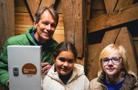 Drie mensen in winterjassen poseren in een houten ruimte, één houdt een kastje met sticker "Beersel". (vergroot foto)