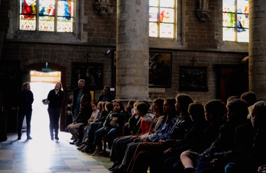 Groep kinderen zit in de kerk van Alsemberg met hoge bogen en kleurrijke glas-in-loodramen. (vergroot foto)