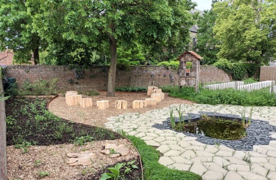 Een groene tuin met een vijver, houten zitblokken in een cirkel en een insectenhotel tegen een bakstenen muur. (vergroot foto)