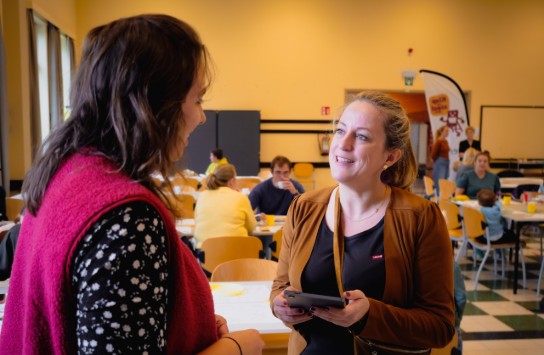 Twee vrouwen praten in een zaal met tafels en stoelen, waar meerdere mensen zitten en staan. (vergroot foto)
