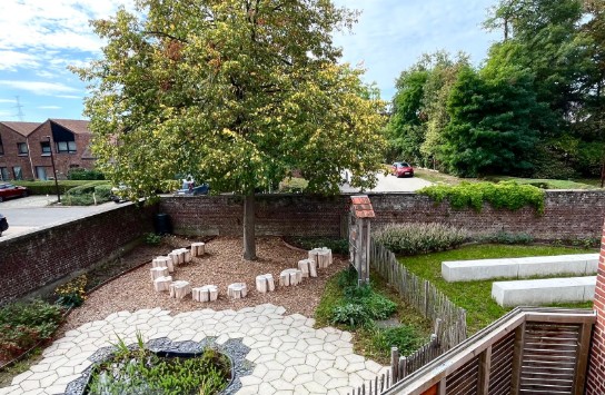 Tuin met boom, houten zitblokken in cirkel, vijver met planten, grasveld en stenen banken, omgeven door bakstenen muur. (vergroot foto)