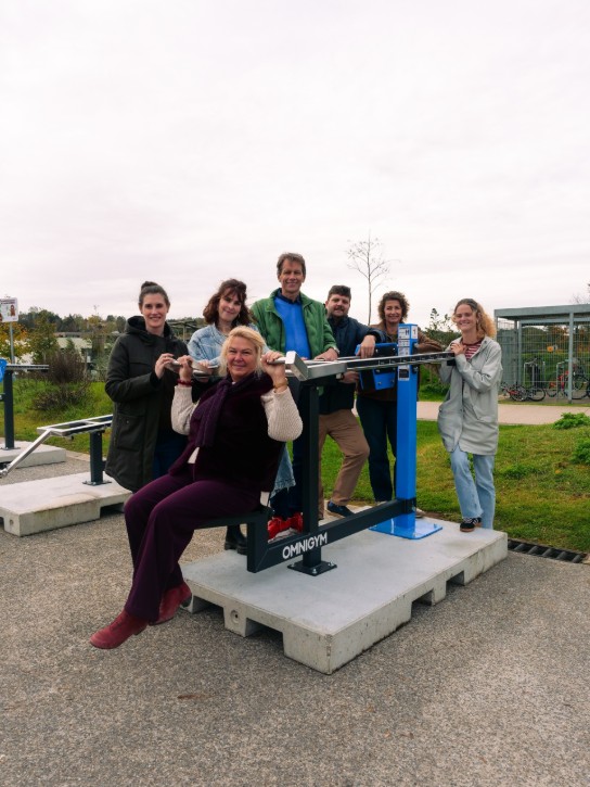 Zeven mensen poseren bij een outdoor fitnessapparaat; één vrouw zit erop. (vergroot foto)