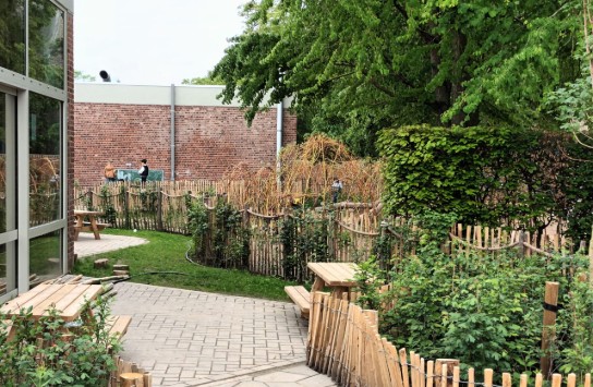 Buitenterrein met houten hekken, bankjes, groen en een bakstenen muur op de achtergrond. (vergroot foto)